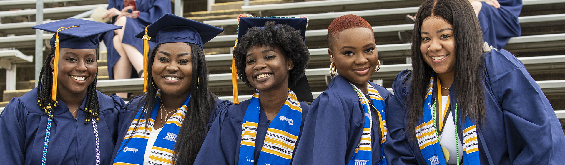 commencement210510a002 Commencement - 5 students in cap and gown smiling and sitting on bleachers