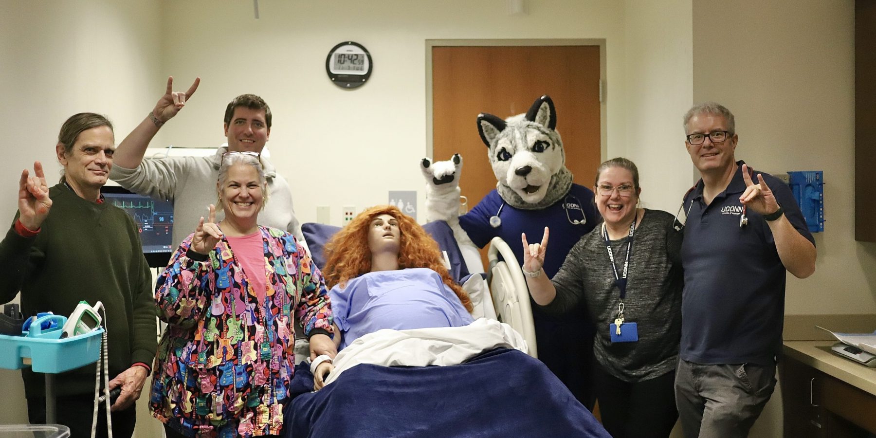 SIM Lab Jonathan 23 Simulation Lab faculty making Husky sign with their hands standing around a manikin and with the mascot Jonathan the Husky