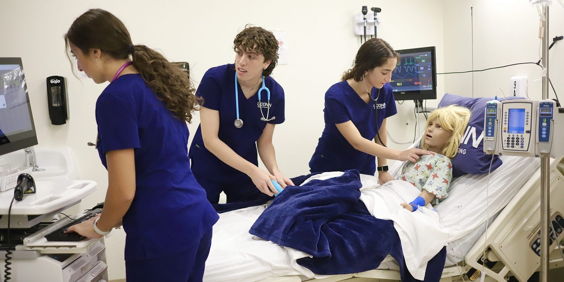 sim lab new 23 Three nursing students in scrubs taking pulse of manikin