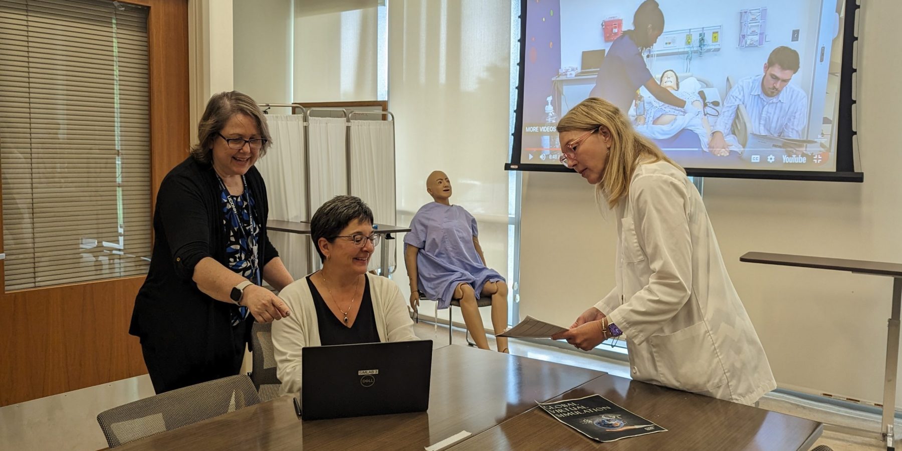 sim2- 2023 Three female faculty smiling and standing behind a laptop in the sim lab